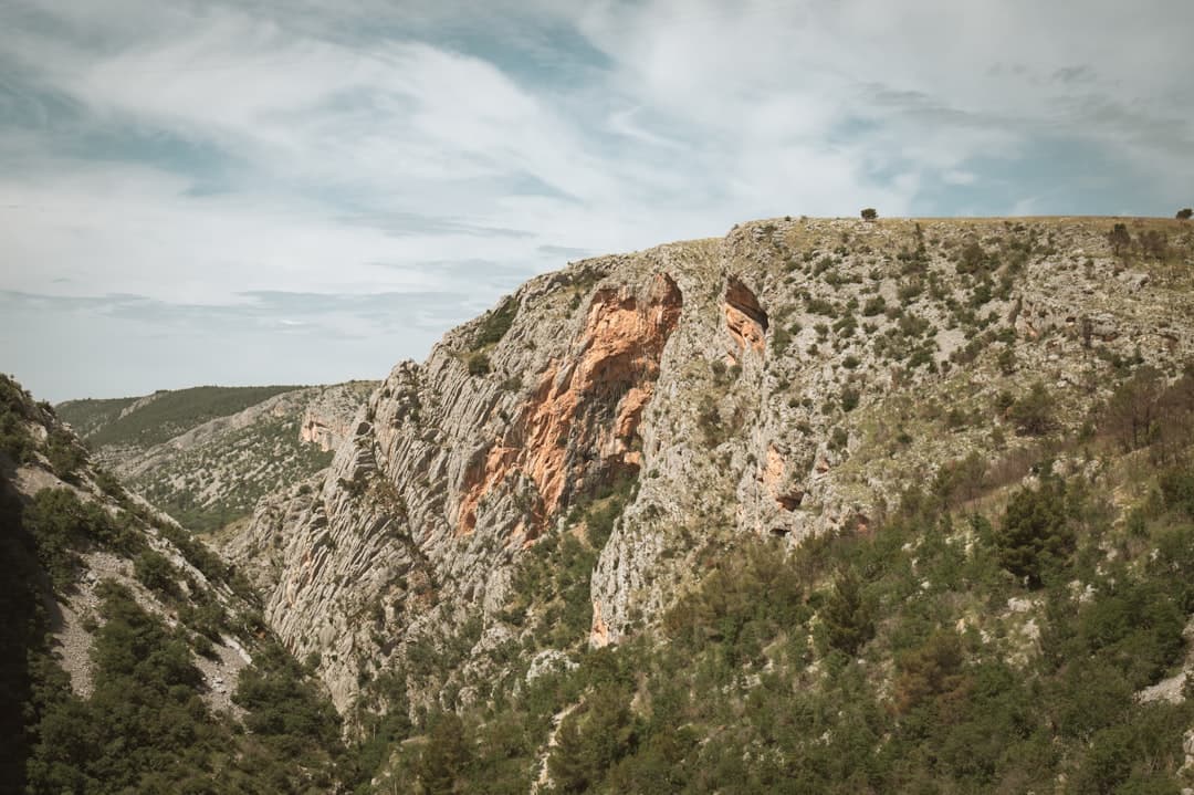 A sheer rock face flanked by pine forest above the canyon floor, evoking the cliffs at Klanci where the Tito tunnels are cut into the wall.