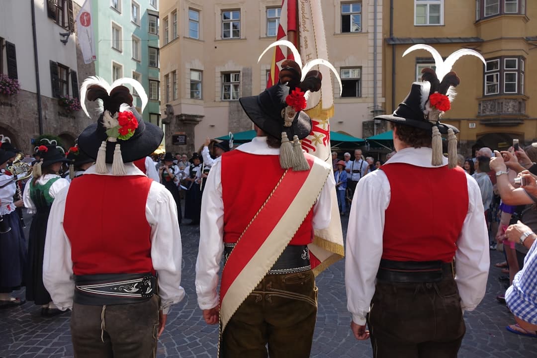 Figuranti folk con panciotto rosso e alti copricapi di piume e fiori, visti di spalle in una piazza cittadina, evocazione della tradizione costumistica dei gruppi carnevaleschi della regione di Kastav.