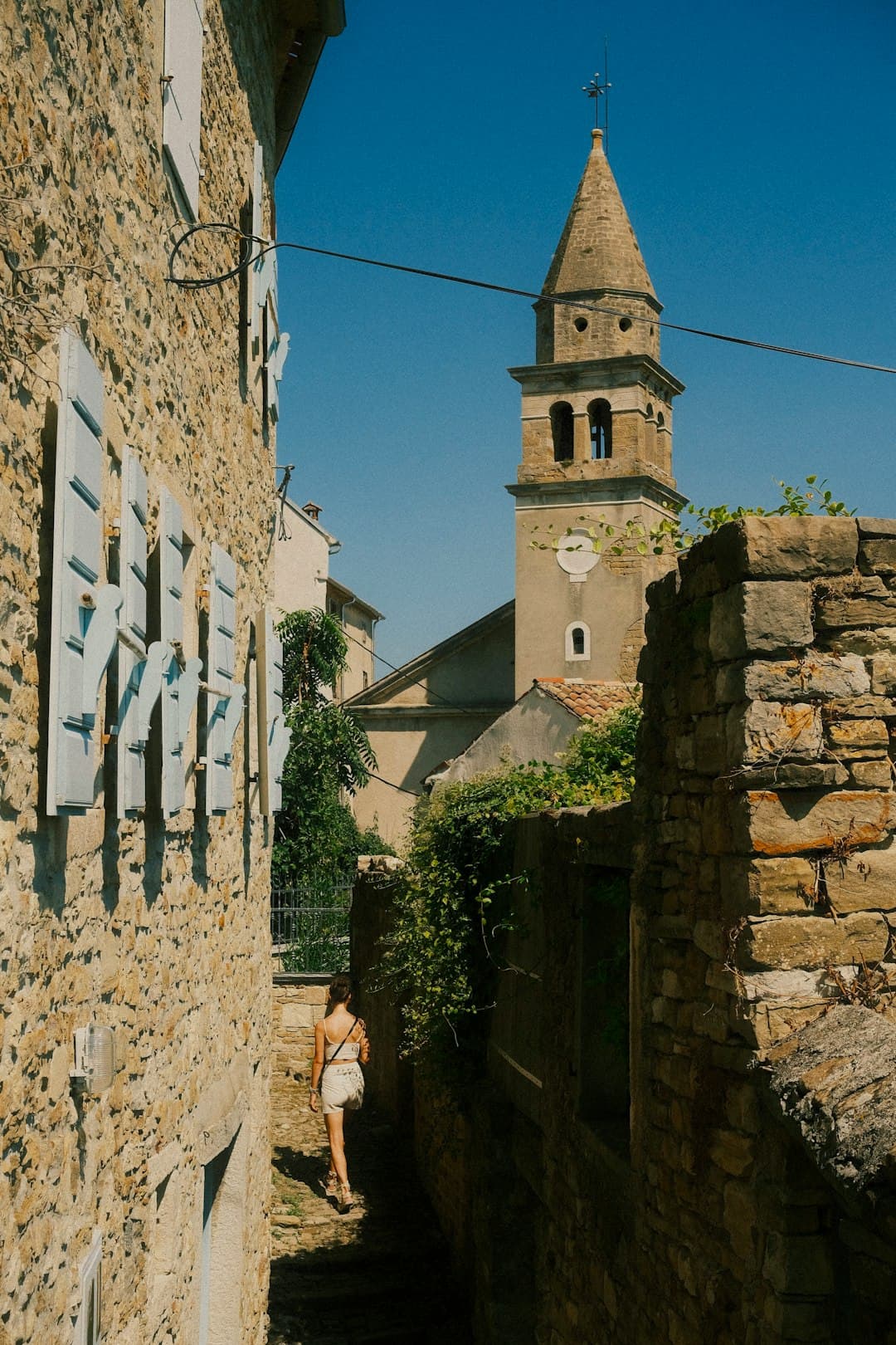 Una donna sale un vicolo selciato di Motovun verso il campanile ottagonale della chiesa, con la luce del tardo pomeriggio sui muri di calcare.
