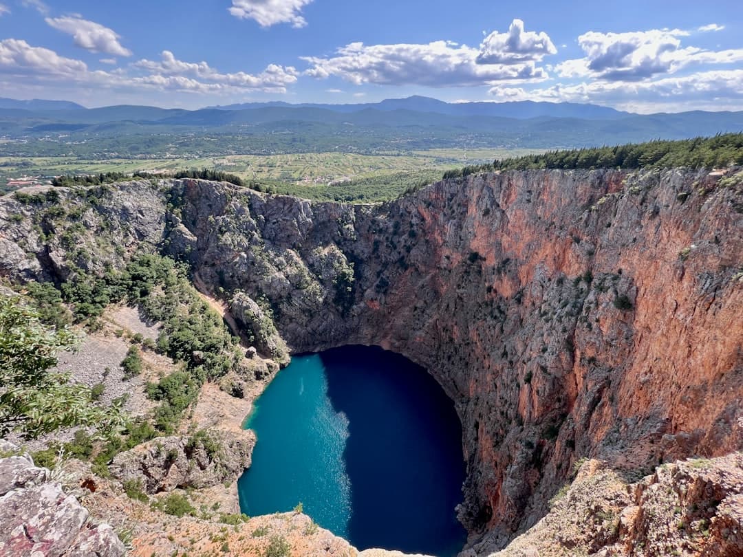 View from the rim down into a karst sinkhole near Imotski, with iron-stained cliffs dropping to a small disc of dark water far below.