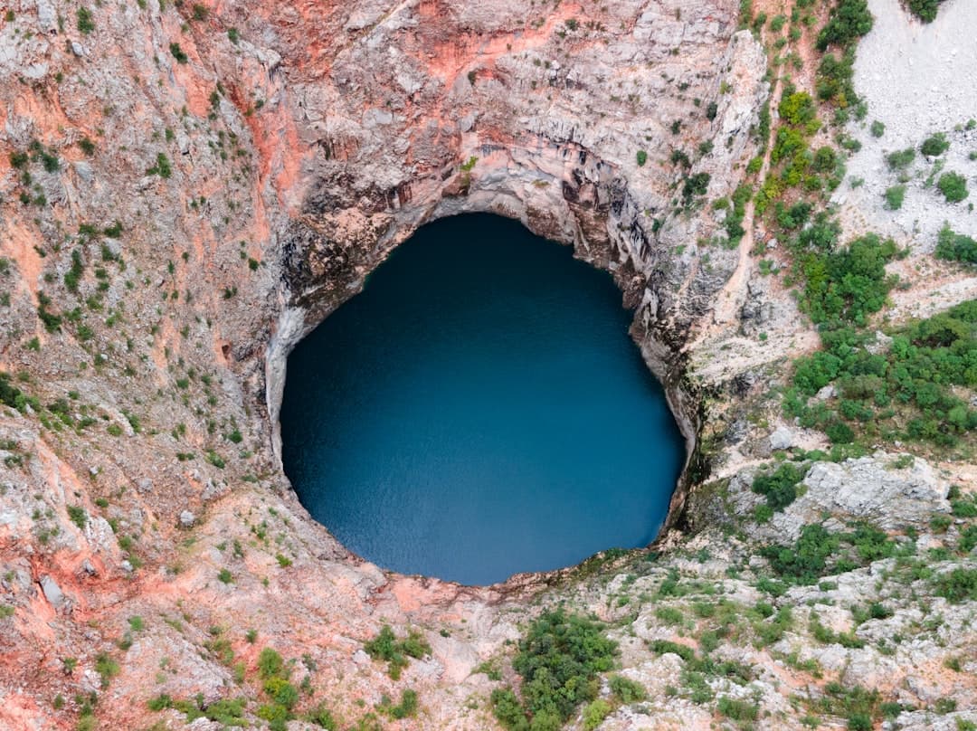 Close framing of the reddish-orange limestone walls of a collapse doline plunging vertically to a still pool of dark blue water.