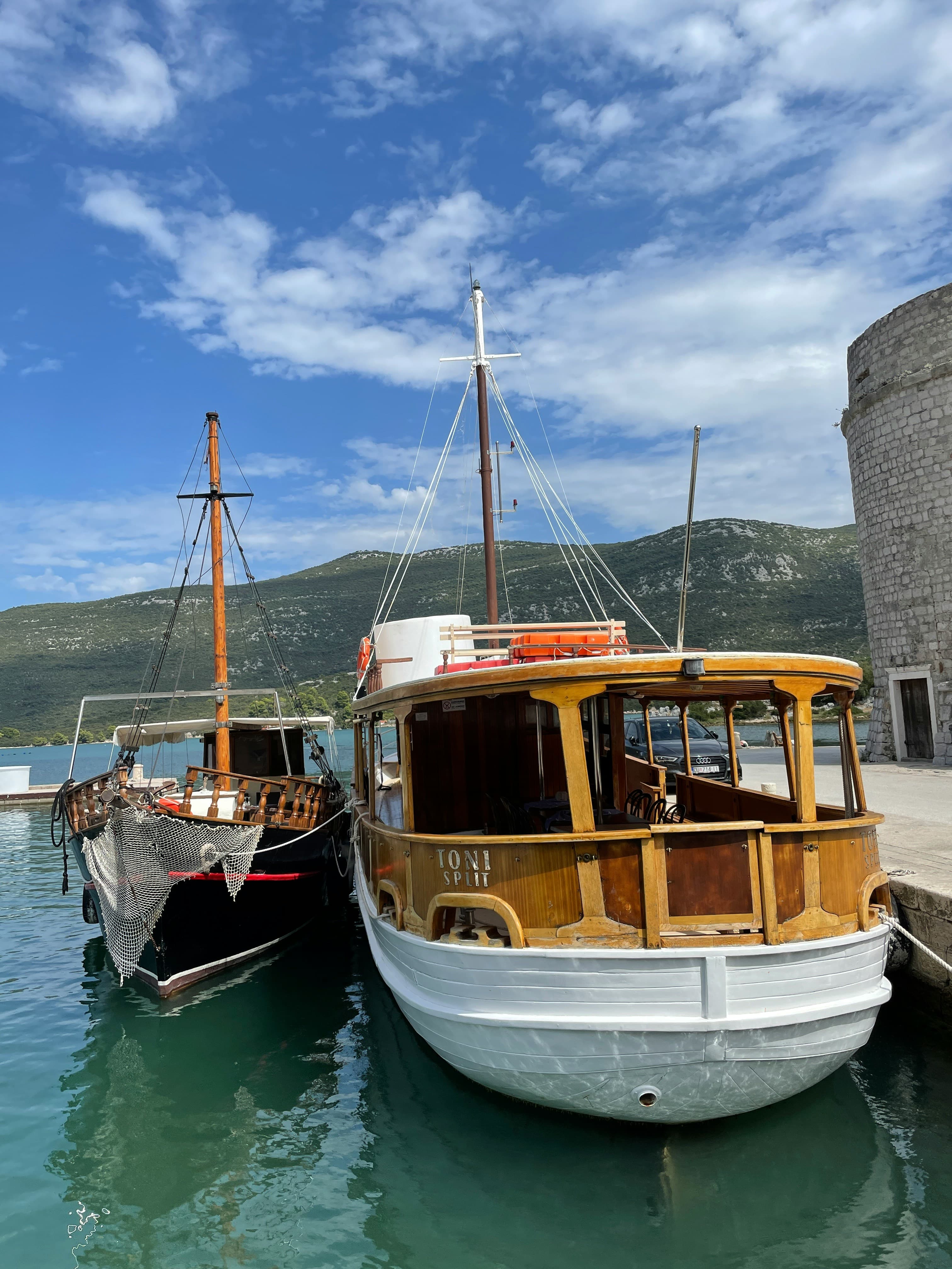 Due barche di legno ormeggiate al porto di Mali Ston, con una torre di pietra rotonda delle Mura di Ston visibile sulla destra.